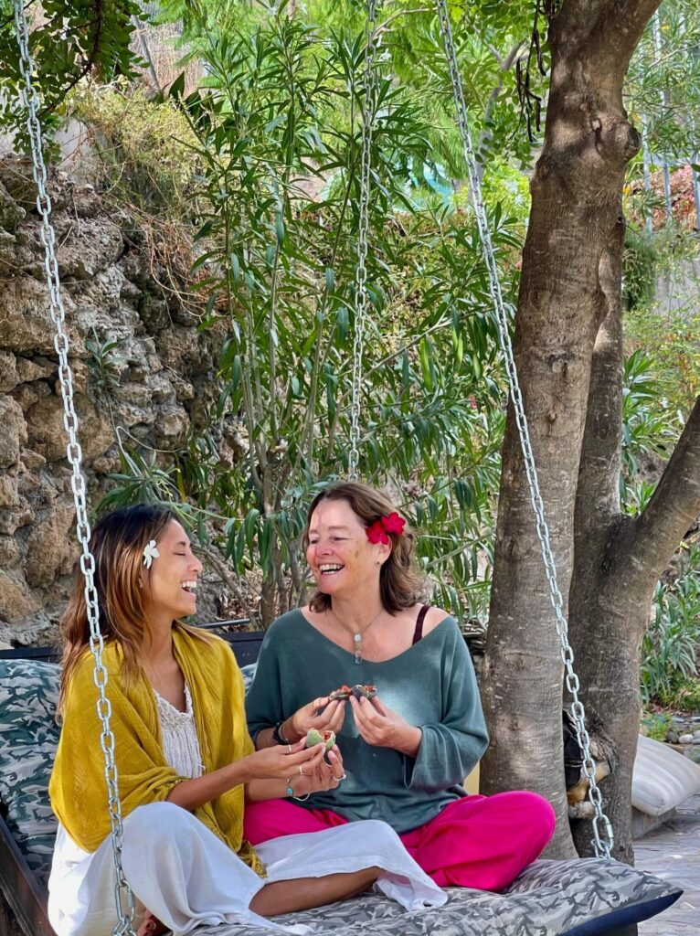 Somarising sisters - Julie Kent and Michelle Kent - smiling with flowers in hair sitting on a garden swing at Molino Del Rey retreat centre Andalusia