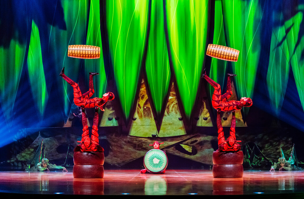 Cirque du Soleil performers in insect-inspired costumes execute a foot-juggling sequence during Cirque du Soleil OVO, set against the show’s vivid green stage design.