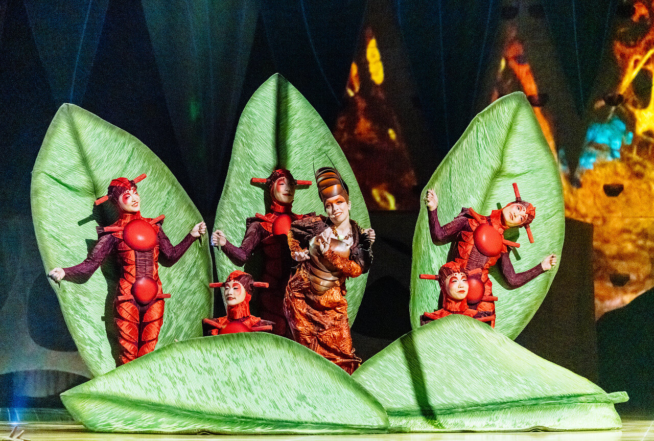 Performers in insect-inspired costumes appear among oversized green leaves during an ensemble scene in Cirque du Soleil OVO.