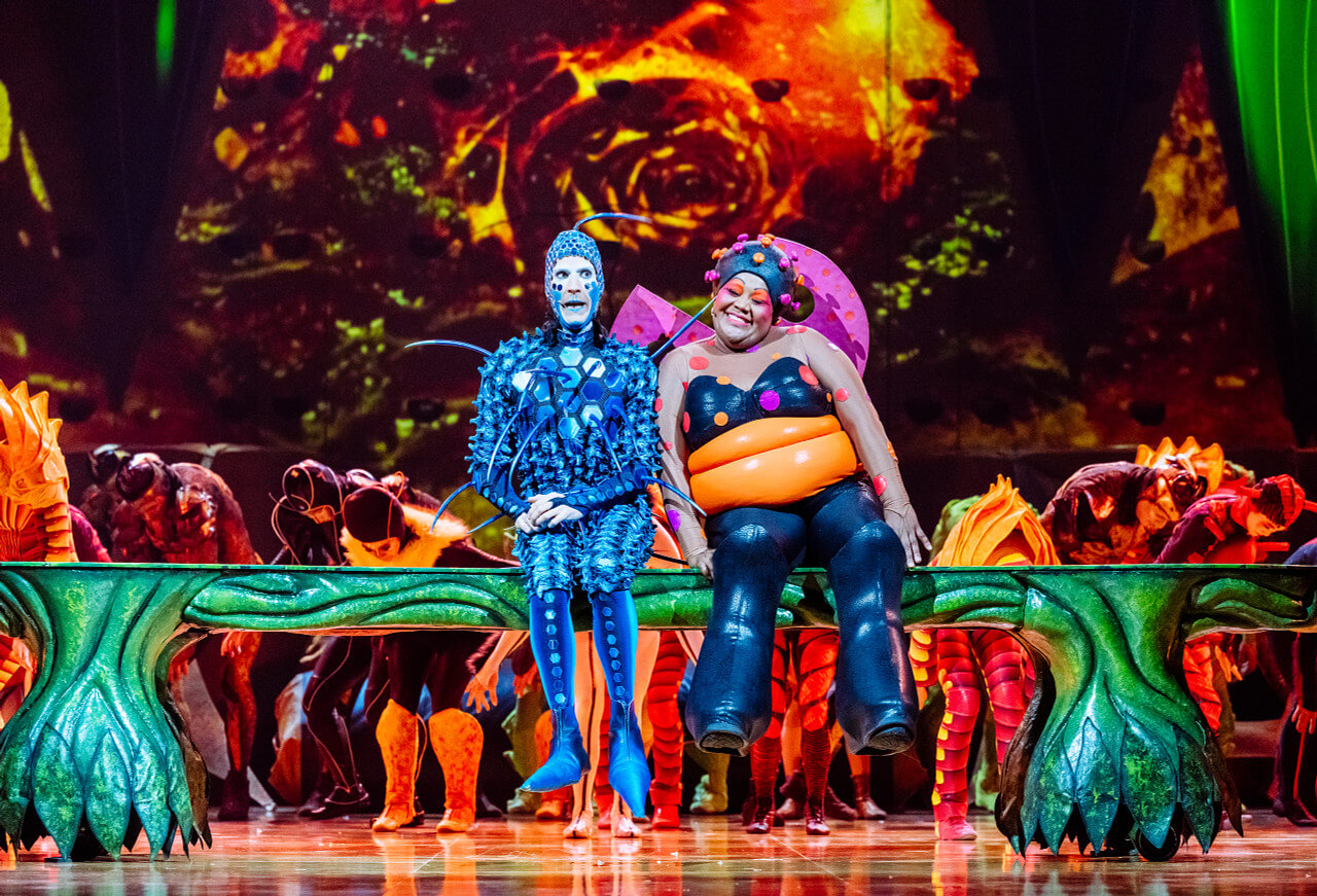 Two lead characters sit together on stage during Cirque du Soleil OVO, depicting the developing love story between the blue fly known as ‘the Foreigner’ and a high-spirited ladybird.